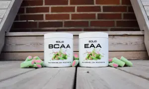 Två burkar, en med BCAA och en med EAA, står framför en tegelvägg. Runt burkarna ligger gröna godisar med rosa fyllning.