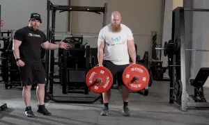 En man med skägg utför farmers walk med tunga vikter. En tränare visar hur man gör.