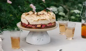 En vacker sockerkaka med rabarber och maräng, serverad utomhus med glas och en flaska dryck.