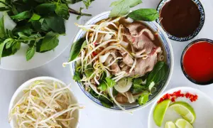 En skål med vietnamesisk mat, pho, med nudlar, tunt skivat kött, böngroddar och färska örter. Tillbehör som lime, chili och såser.