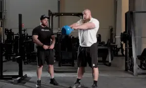 Två män i gym, en utför kettlebell swings med en blå kettlebell.