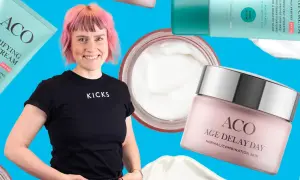 En kvinna med rosa hår ler omgiven av ACO-produkter, inklusive ACO ansiktskräm anti age.