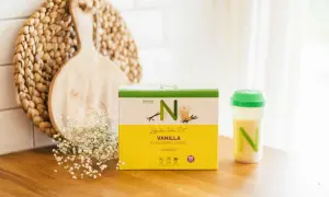 En låda med Nutrilett vaniljshake för en vlcd diet står på en träbänk. Bredvid finns en shaker med shake och en dekoration av trä och blommor.