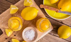 Inlagda citroner i glasburkar, färska citroner, salt och pepparkorn på en träskärbräda.