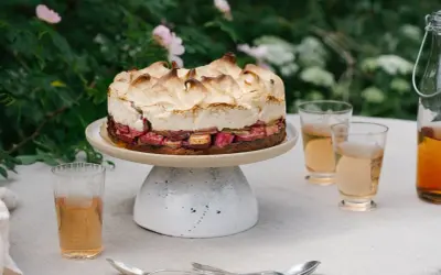 En vacker sockerkaka med rabarber och maräng, serverad utomhus med glas och en flaska dryck.