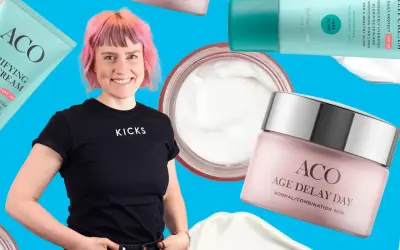 En kvinna med rosa hår ler omgiven av ACO-produkter, inklusive ACO ansiktskräm anti age.