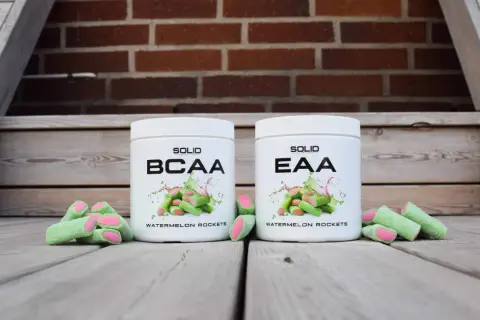 Två burkar, en med BCAA och en med EAA, står framför en tegelvägg. Runt burkarna ligger gröna godisar med rosa fyllning.