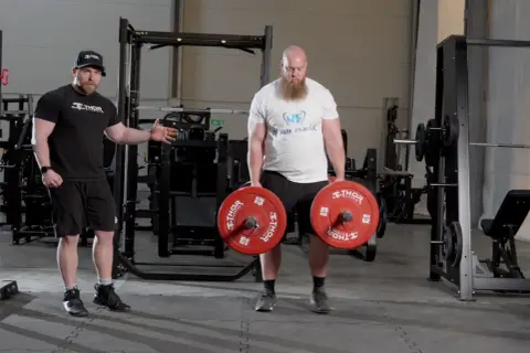 En man med skägg utför farmers walk med tunga vikter. En tränare visar hur man gör.
