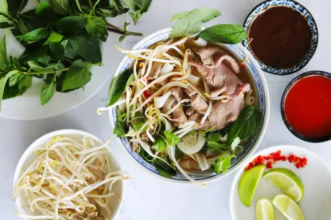 En skål med vietnamesisk mat, pho, med nudlar, tunt skivat kött, böngroddar och färska örter. Tillbehör som lime, chili och såser.