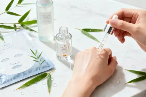 En hand applicerar C-vitaminserum för j beauty. Bambublad och sheet mask i bakgrunden.