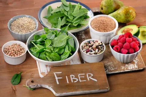 En skärbräda med texten "FIBER" omgiven av skålar med bönor, linser, spenat, sockerärtor, hallon, avokado, päron och korn. En hyllning till fiberrik mat.