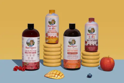 Flera flaskor Mary Ruths multivitamin i olika smaker, inklusive barnvarianten. Frukt som hallon, mango och äpple finns bredvid.