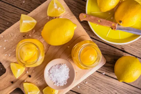 Inlagda citroner i glasburkar, färska citroner, salt och pepparkorn på en träskärbräda.