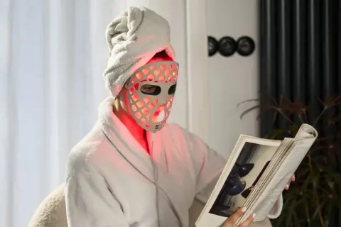Person med handduk på huvudet och en led mask ansikte läser en bok i en morgonrock.