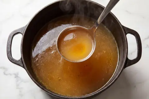 En slev lyfter gyllene, ångande buljong från en gjutjärnsgryta. Doften av real broth savory spices sprider sig.
