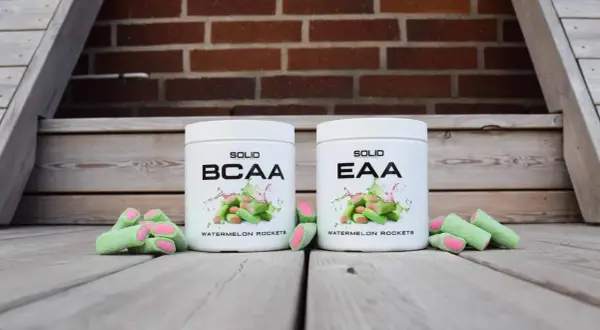 Två burkar, en med BCAA och en med EAA, står framför en tegelvägg. Runt burkarna ligger gröna godisar med rosa fyllning.