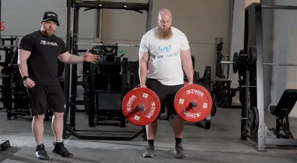 En man med skägg utför farmers walk med tunga vikter. En tränare visar hur man gör.