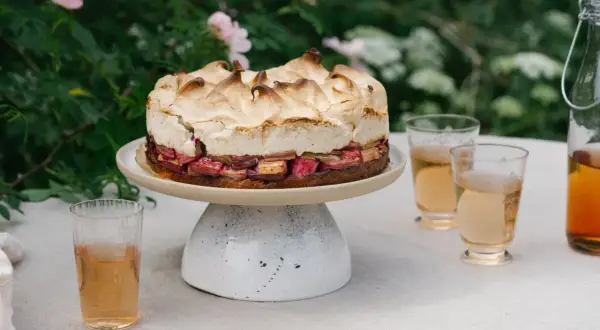 En vacker sockerkaka med rabarber och maräng, serverad utomhus med glas och en flaska dryck.