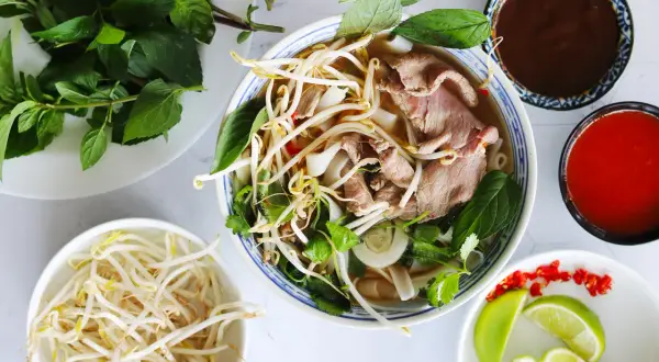 En skål med vietnamesisk mat, pho, med nudlar, tunt skivat kött, böngroddar och färska örter. Tillbehör som lime, chili och såser.