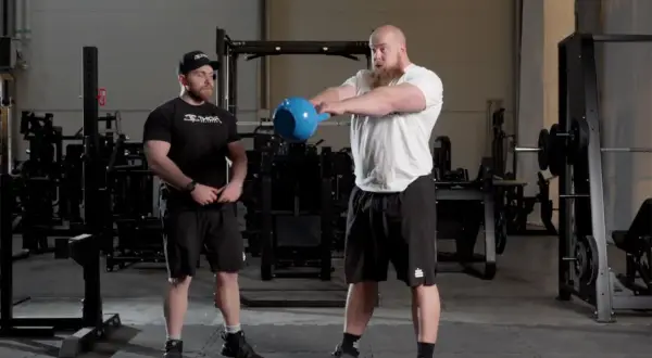 Två män i gym, en utför kettlebell swings med en blå kettlebell.