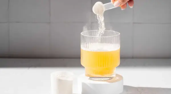 En hand häller pulver från en skopa ner i ett glas med gul vätska. Pulvret är en del av matters multi collagen.