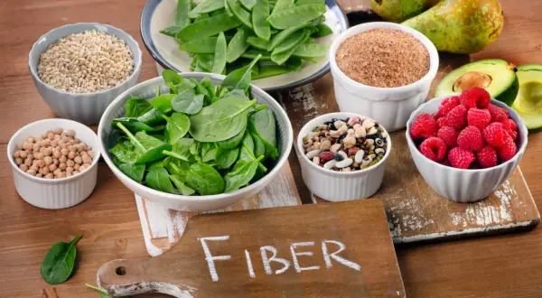 En skärbräda med texten "FIBER" omgiven av skålar med bönor, linser, spenat, sockerärtor, hallon, avokado, päron och korn. En hyllning till fiberrik mat.