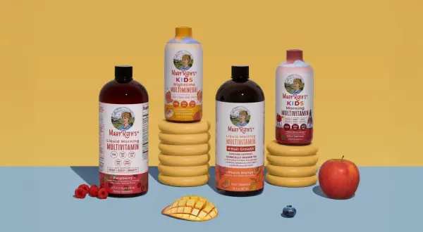 Flera flaskor Mary Ruths multivitamin i olika smaker, inklusive barnvarianten. Frukt som hallon, mango och äpple finns bredvid.