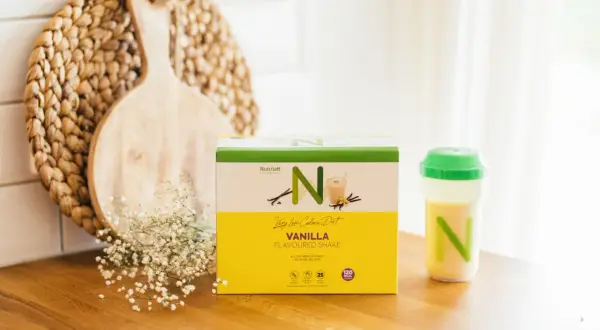 En låda med Nutrilett vaniljshake för en vlcd diet står på en träbänk. Bredvid finns en shaker med shake och en dekoration av trä och blommor.