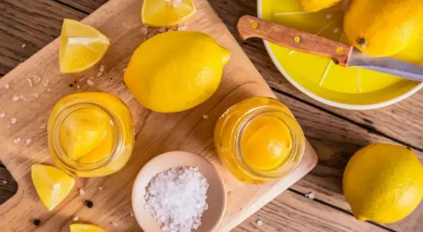Inlagda citroner i glasburkar, färska citroner, salt och pepparkorn på en träskärbräda.