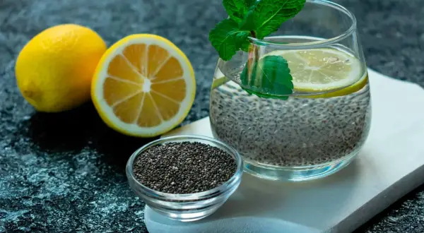 Ett glas med chia vatten, garnerat med citron och mynta. Bredvid står en skål med chiafrön och två citroner.