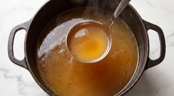 En slev lyfter gyllene, ångande buljong från en gjutjärnsgryta. Doften av real broth savory spices sprider sig.