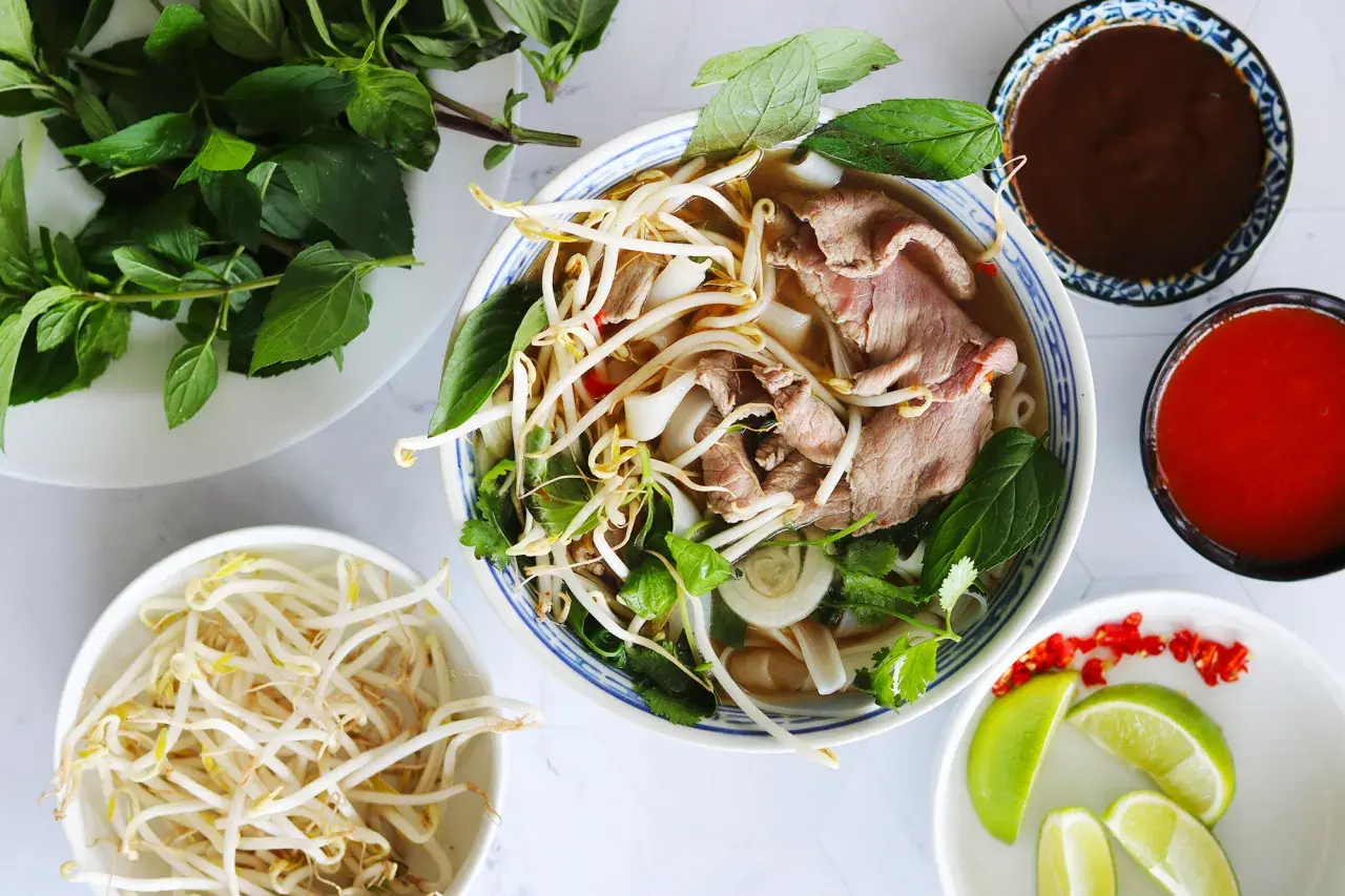 En skål med vietnamesisk mat, pho, med nudlar, tunt skivat kött, böngroddar och färska örter. Tillbehör som lime, chili och såser.
