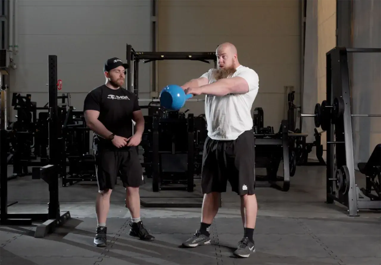 Två män i gym, en utför kettlebell swings med en blå kettlebell.