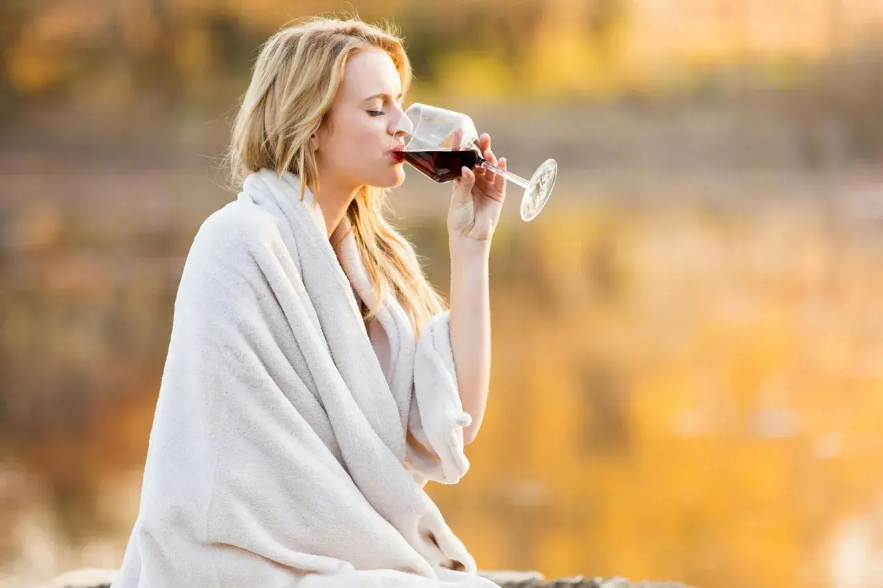 Kvinna med blont hår dricker rödvin insvept i en filt. Visste du att kalorier vitt vin per dl kan variera?