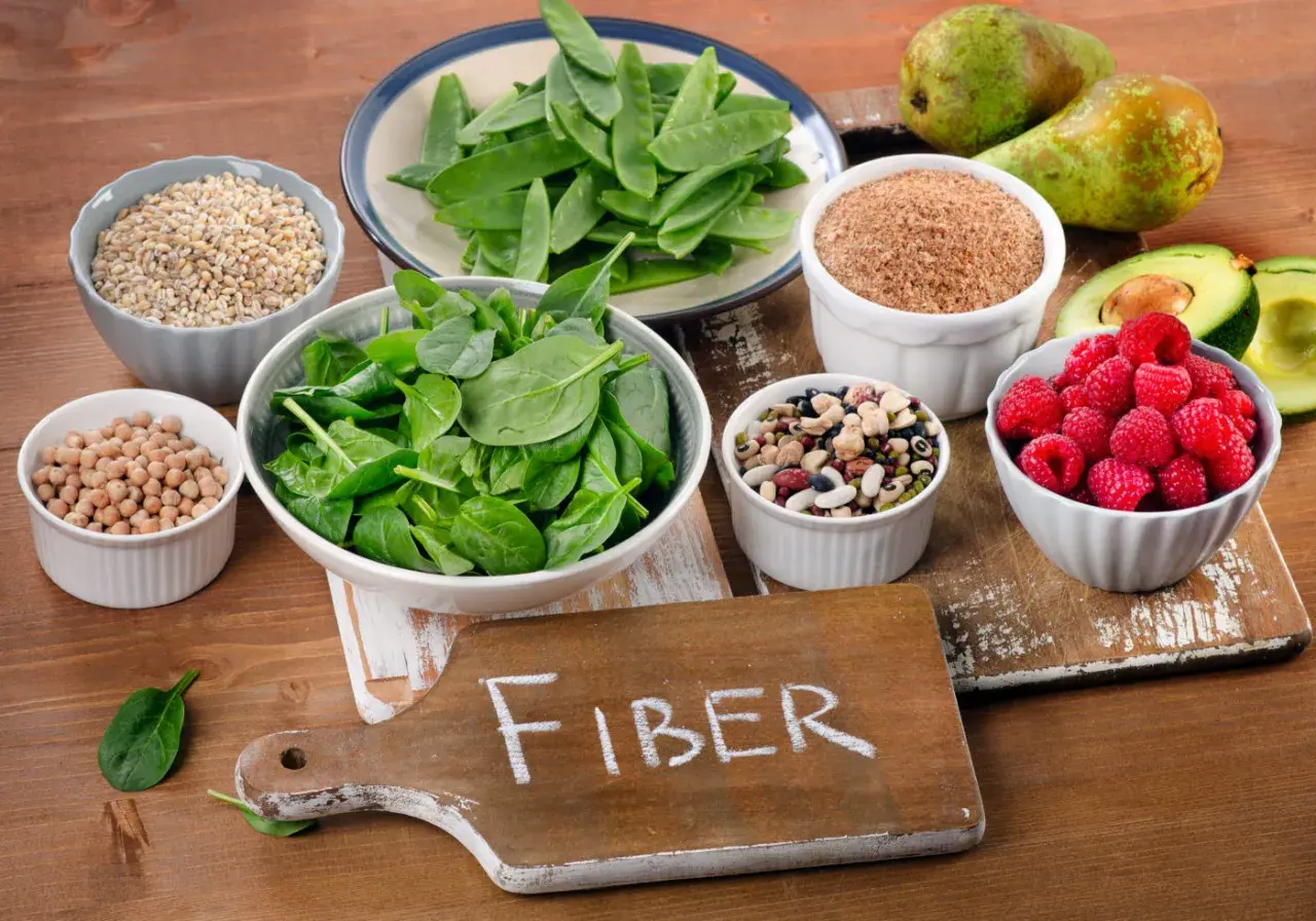 En skärbräda med texten "FIBER" omgiven av skålar med bönor, linser, spenat, sockerärtor, hallon, avokado, päron och korn. En hyllning till fiberrik mat.