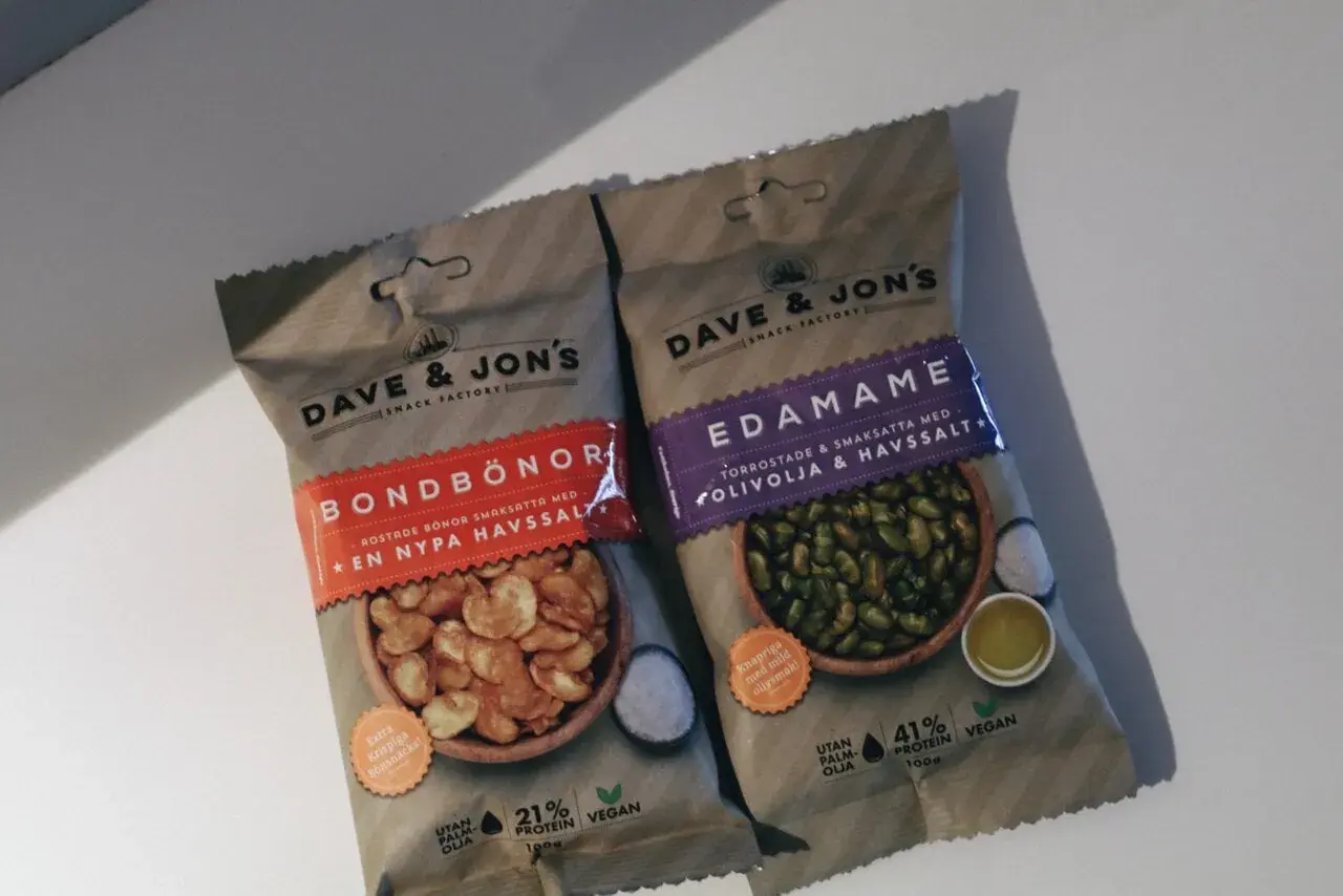 Två påsar Dave & Jon's snacks: Bondbönor och Edamame. Edamame-påsen visar torrostade edamamebönor med olivolja och havssalt.
