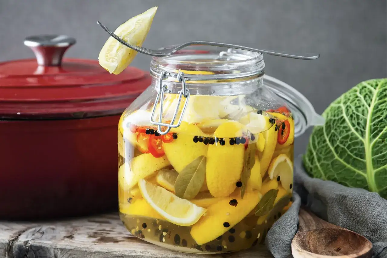 En stor glasburk fylld med inlagda citroner, chili, pepparkorn och lagerblad. En gaffel med en citronbit sticker upp ur burken.