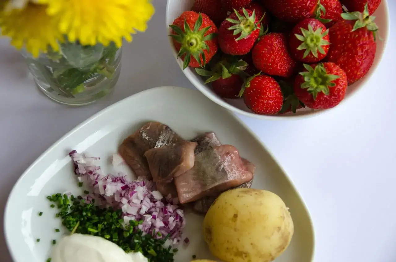 F&ouml;rberedelser f&ouml;r en midsommar buffe: sill, potatis, gr&auml;ddfil, gr&auml;sl&ouml;k, r&ouml;dl&ouml;k och jordgubbar. En gul blomma i bakgrunden.