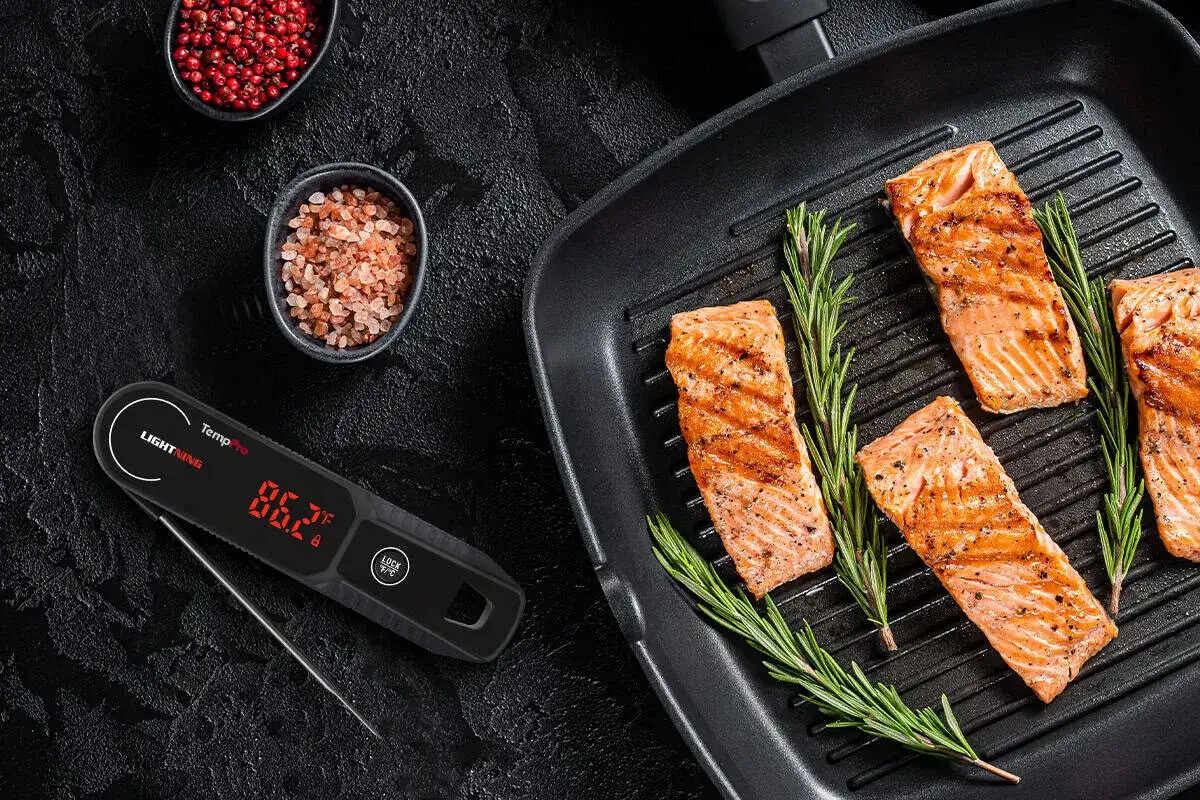 Grillad lax med rosmarin i en grillpanna, bredvid en digital termometer som visar lax innertemperatur. Sm&aring; sk&aring;lar med pepparkorn och flingsalt.