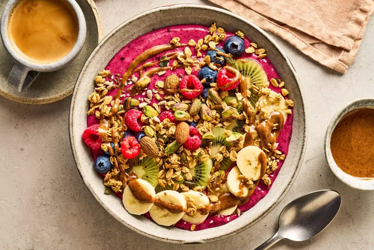 En f&auml;rgglad smoothie bowl med b&auml;r, frukt, n&ouml;tter och granola. Perfekt f&ouml;r en h&auml;lsosam 5:2 frukost. Bredvid en kopp kaffe och en sk&aring;l med n&ouml;tsm&ouml;r.