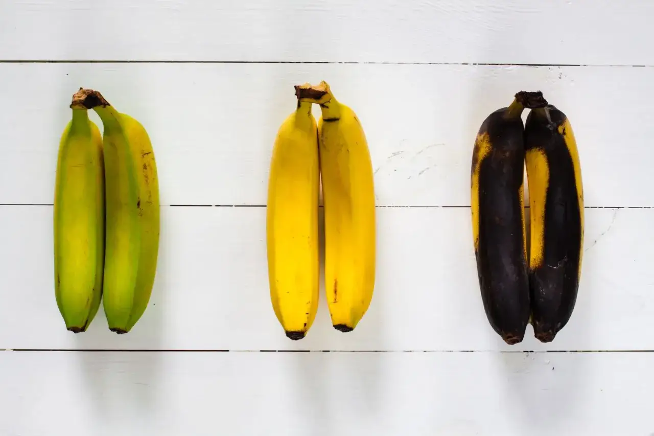 Tre par bananer på vit träbakgrund: gröna, gula och övermogna. Banan nyttigt i alla stadier, men bäst när de är gula.
