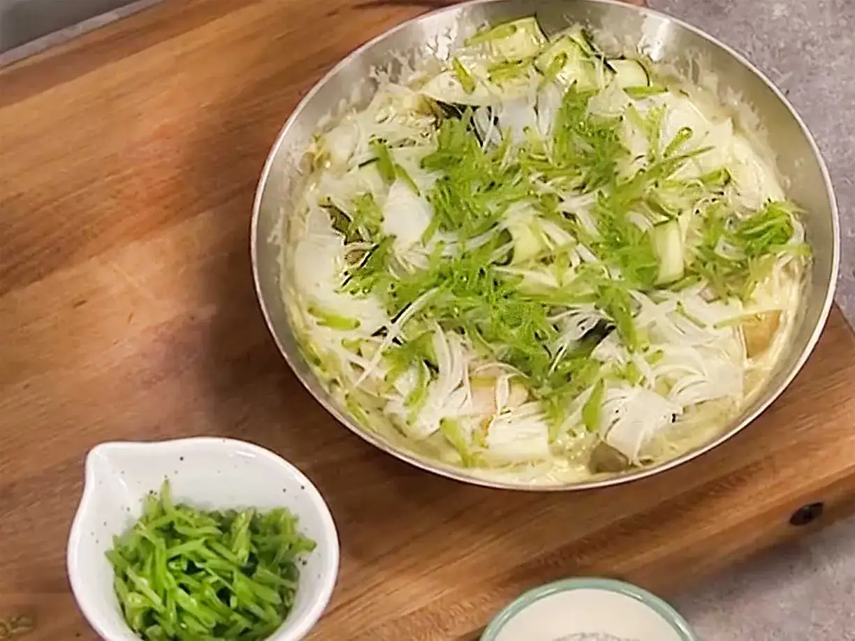 En panna med vit fisk och gr&ouml;nsaker, toppad med strimlad gr&ouml;nk&aring;l. En liten sk&aring;l med gr&ouml;na strimlor st&aring;r bredvid.