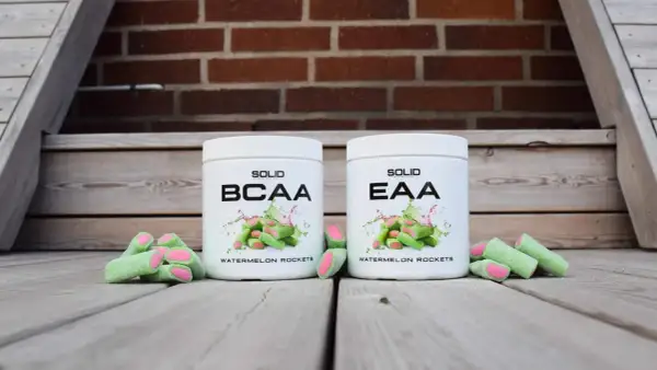Två burkar, en med BCAA och en med EAA, står framför en tegelvägg. Runt burkarna ligger gröna godisar med rosa fyllning.