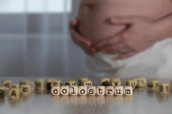 Gravid mage med händerna vilande på magen. Träpärlor med bokstäver bildar ordet "colostrum svenska", som beskriver den första mjölken.