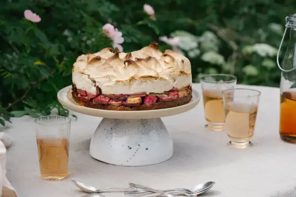 En vacker sockerkaka med rabarber och maräng, serverad utomhus med glas och en flaska dryck.