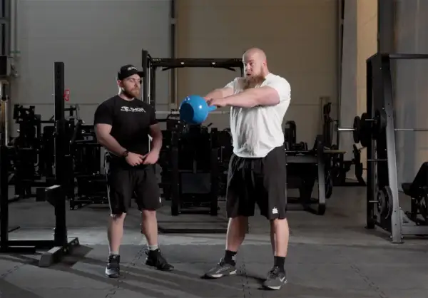 Två män i gym, en utför kettlebell swings med en blå kettlebell.