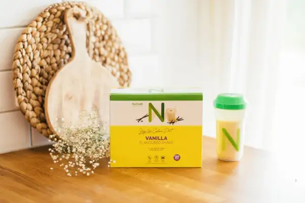 En låda med Nutrilett vaniljshake för en vlcd diet står på en träbänk. Bredvid finns en shaker med shake och en dekoration av trä och blommor.